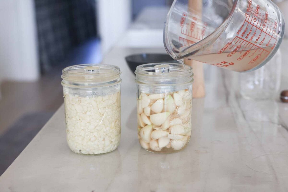 Garlic in mason jars with a weight on them and a salt brine being poured into the jars.
