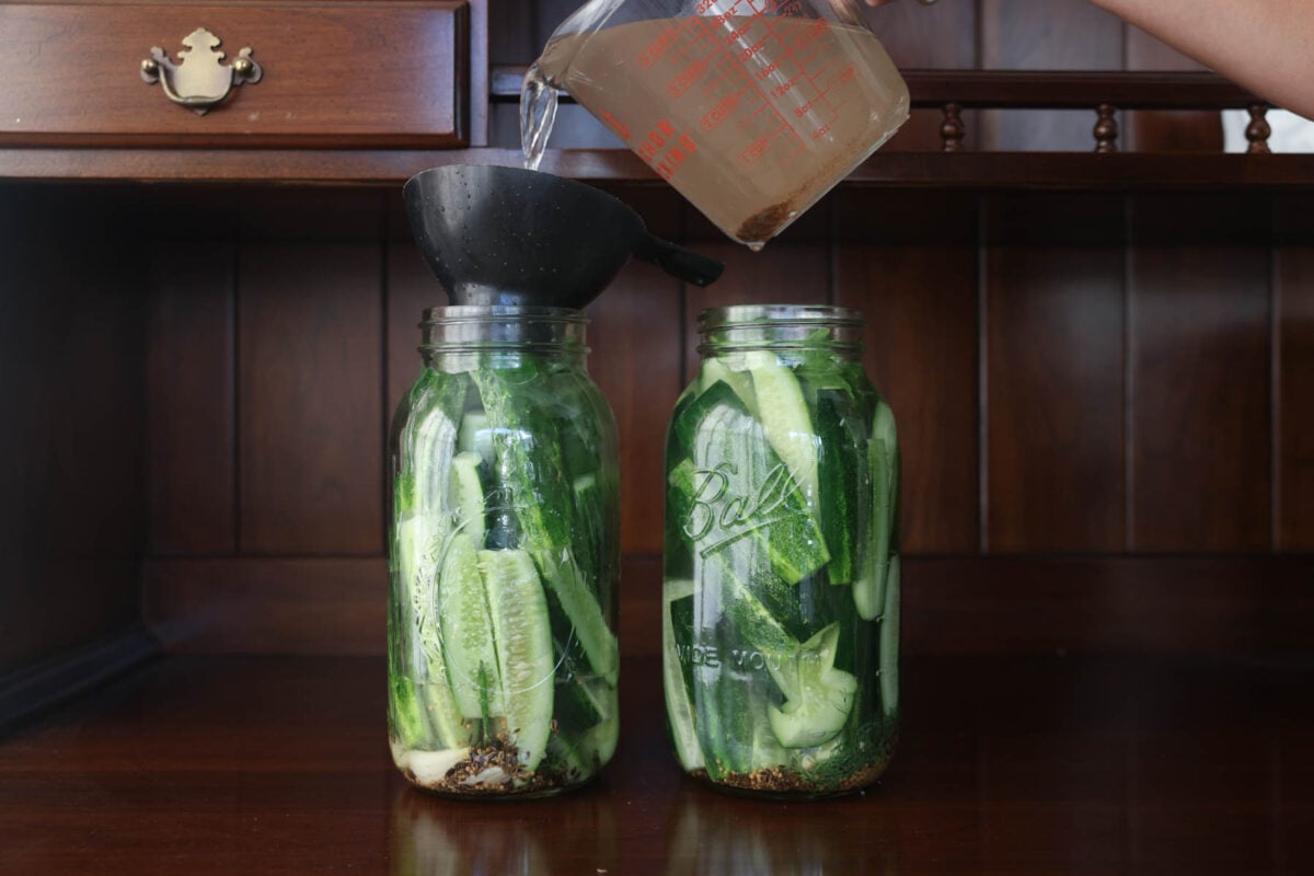Pouring a brine into mason jars with cucumbers, dill, and spices.