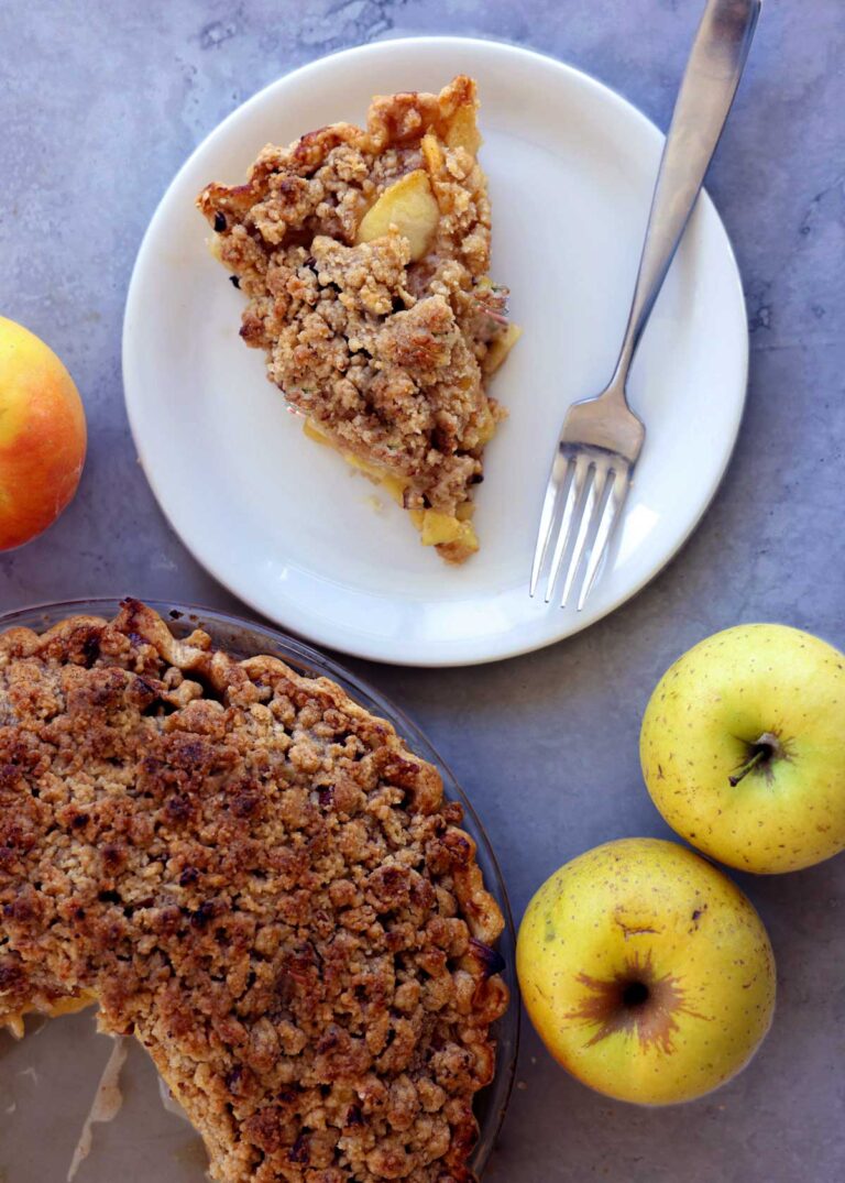 A slice of dutch apple pie on a place with the remaining pie on the table along with some apples.
