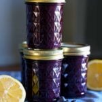 A couple jars of blueberry lemon jam stacked on each other with lemons on the table nearby.