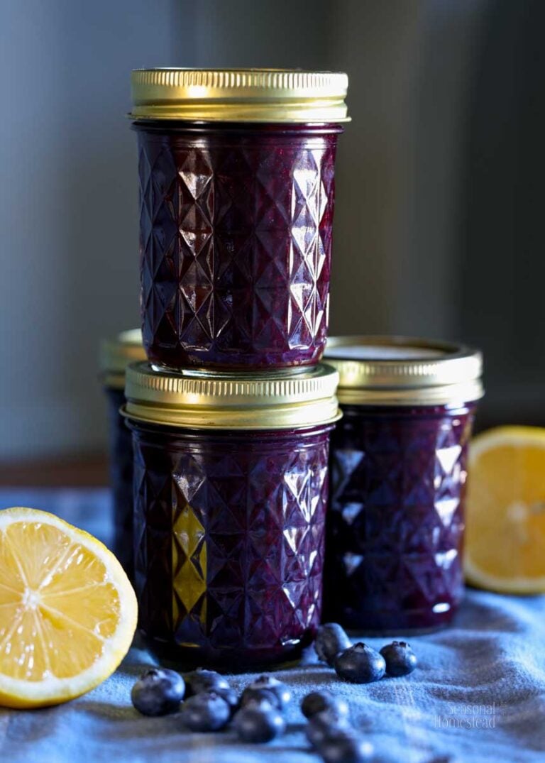 A couple jars of blueberry lemon jam stacked on each other with lemons on the table nearby.