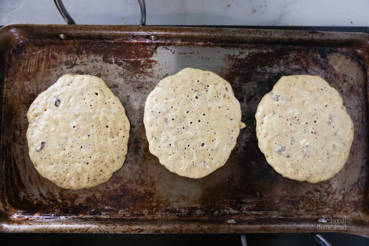 Multigrain pancakes are partially cooked on a griddle showing bubbles on top.