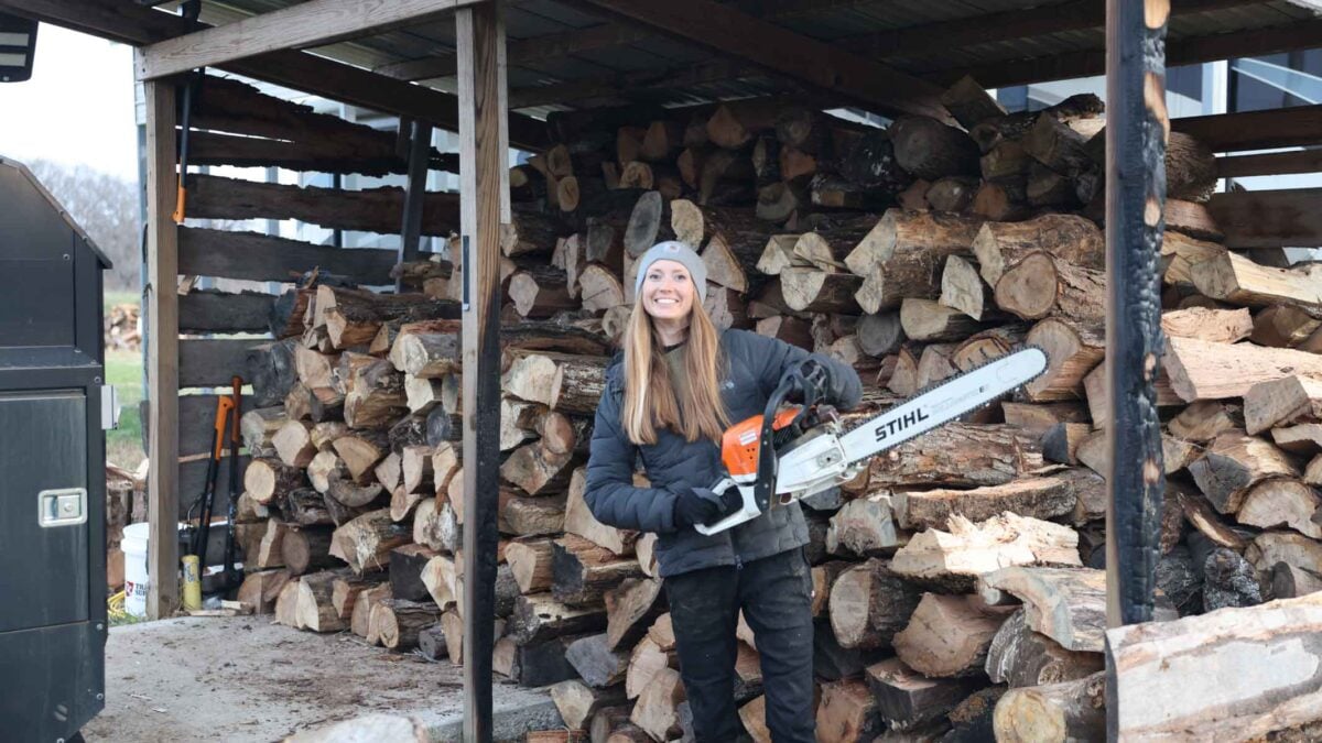 Becky stands next to a large outdoor woodpile with a chainsaw.