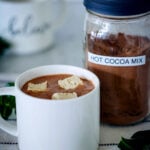 hot cocoa in a cup with marshmallows and hot cocoa mix in a jar behind it.